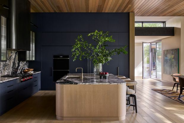 A sophisticated open-plan kitchen with a large rounded island clad in light timber cabinetry and topped with dramatic veined dark stone. Deep navy built-in cabinetry and matching dark marble backsplash create a bold focal wall, while integrated appliances and a brass-toned tap add refined detailing. A warm timber-lined ceiling continues into the adjoining dining and living area, which is visible beyond the kitchen. Full-height glazing and an open layout bring in abundant natural light.