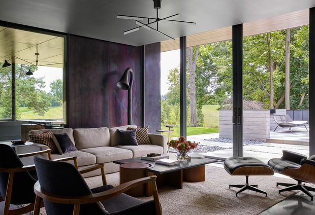A sophisticated contemporary living room with floor-to-ceiling glazing opening onto a landscaped outdoor terrace. The room features a large neutral sectional sofa, mid-century-inspired lounge chairs, a black leather recliner and ottoman, sculptural floor lamp, low coffee tables, and a dramatic dark textured feature wall. A linear modern pendant hangs from the ceiling, while the glazing provides views of mature trees and lawn. The space has a refined, warm, indoor-outdoor character.