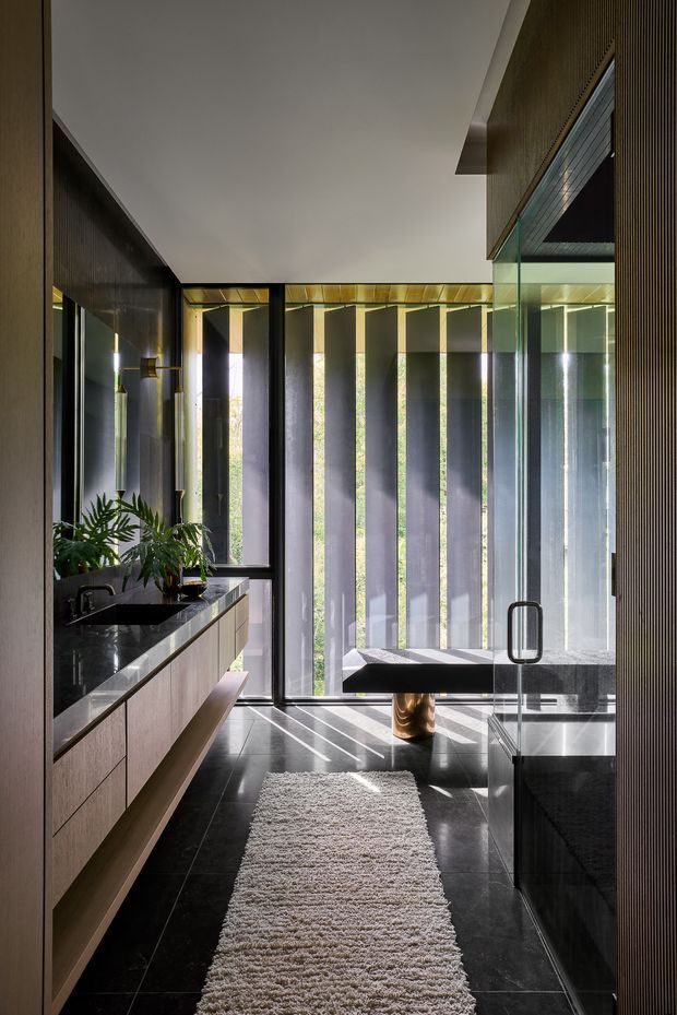 A refined contemporary bathroom with a long floating timber vanity, dark polished stone benchtop and integrated basin, black tapware, and a freestanding bathtub positioned beside full-height glazing. Vertical screening, dark tiled flooring, timber slatted wall detailing, and a textured runner create a dramatic, private atmosphere with strong linear shadows and abundant filtered natural light.