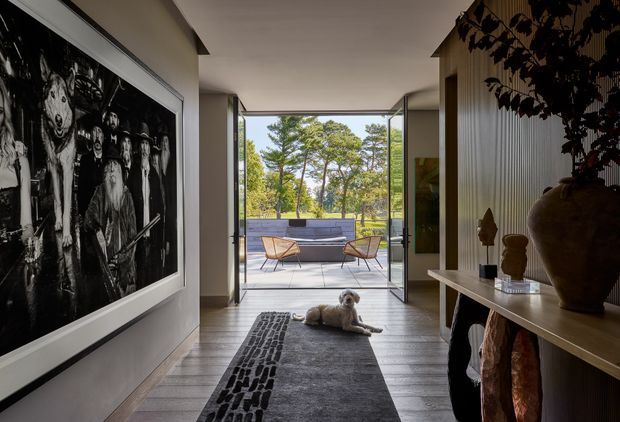 A dark, refined interior hallway or entry passage opens through fully glazed doors to a sunny outdoor terrace and landscaped setting. The space features dark timber flooring, neutral walls, a large monochrome artwork, a long console with sculptural decor, vertical wall panelling, and a patterned charcoal runner. Mid-century style outdoor seating is visible beyond the opening, with a small dog on the rug.