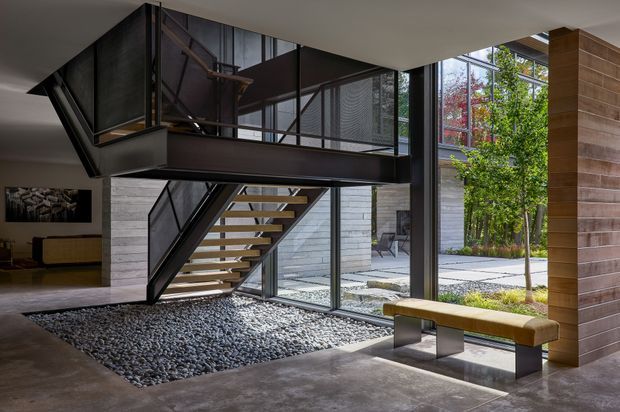 A contemporary open-plan interior centered on a dramatic floating steel-and-timber staircase and mezzanine. Full-height glazing opens onto a landscaped courtyard, while a polished concrete floor, exposed dark structural elements, grey masonry walls, timber wall cladding, and a built-in timber bench create a refined indoor-outdoor composition. A pebble-filled stair void adds texture and separates the entry zone from the living area.