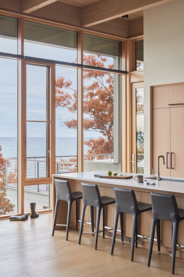 A contemporary open-plan kitchen with a long island and four dark bar stools, positioned beside expansive floor-to-ceiling timber-framed glazing overlooking the ocean. The space features warm timber ceiling beams and flooring, pale fixed cabinetry, a light benchtop, and a stainless steel tap. A balcony and autumn trees are visible beyond the glass.