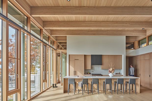 A spacious contemporary kitchen and living zone with a long island bench and multiple stools, integrated timber cabinetry, stainless-steel appliances, and extensive floor-to-ceiling glazing along one side. The room features exposed timber ceiling beams and timber lining, warm timber flooring, and a light, airy connection to the outdoors.