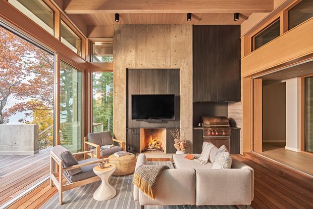 A warm, refined living room with extensive floor-to-ceiling glazing opening onto a timber deck and leafy landscape. The room features a sculptural concrete fireplace wall with integrated television, a wood-burning fire, dark built-in cabinetry, exposed timber ceiling beams, timber-framed glazing, a built-in barbecue or kitchen appliance niche, and contemporary lounge furniture arranged around the fireplace.