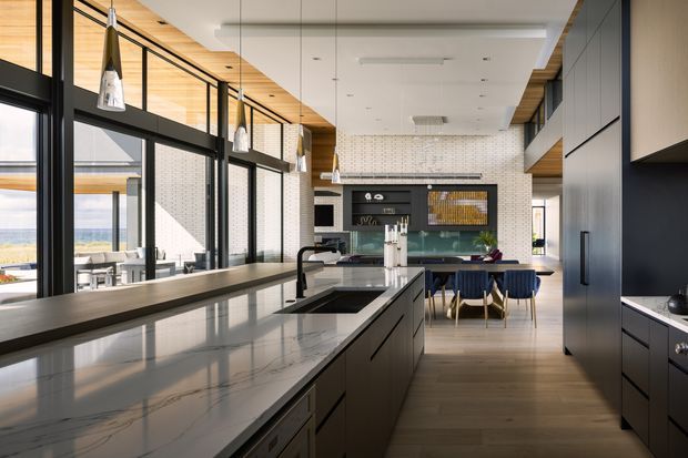 A spacious contemporary kitchen viewed along a long central island toward a dining area. The room features dark flat-panel cabinetry, a light stone island benchtop, large black-framed windows and doors, warm timber ceiling lining, pendant lights, and timber flooring. A tiled backsplash and integrated cooking zone sit at the far end, with abundant natural light and an outdoor outlook.