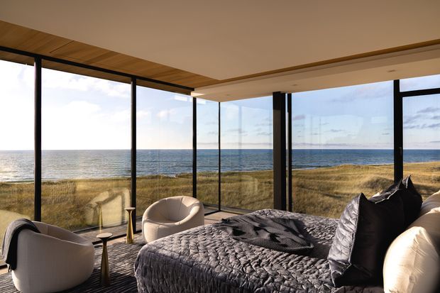 A serene contemporary bedroom with a large bed in the foreground and near floor-to-ceiling corner glazing overlooking a coastal shoreline and ocean. The room features a dark quilted bedspread, pale upholstered lounge chairs, slender side tables, black-framed glazing, and a warm timber ceiling and deck-like floor, creating an airy yet refined coastal retreat.