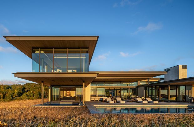A contemporary low-slung residential building with broad cantilevered flat roof planes, extensive floor-to-ceiling glazing, warm timber soffits and cladding, and a large swimming pool in the foreground. The home sits beside landscaped grasses and mature vegetation under a clear blue sky.