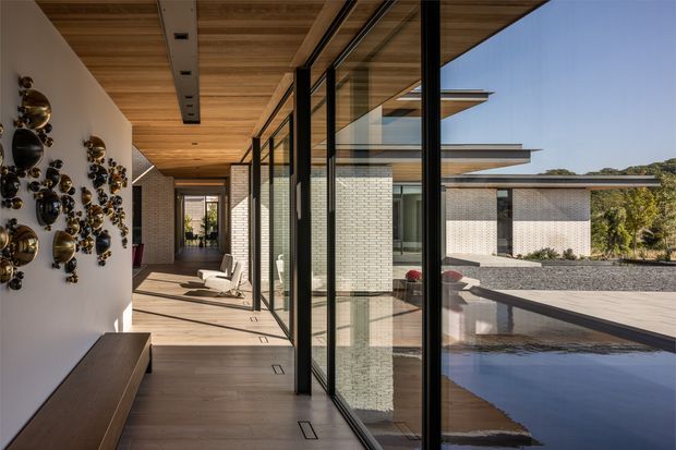 A refined contemporary indoor-outdoor corridor or covered terrace with full-height black-framed glazing overlooking a shallow reflecting pool and a low, flat-roofed brick building. The covered walkway has warm timber ceiling and flooring, pale brick walls, built-in seating, and a decorative sculptural wall installation. Strong visual connections between the interior circulation space, courtyard water feature, and landscaped surroundings create a calm, luxurious atmosphere.