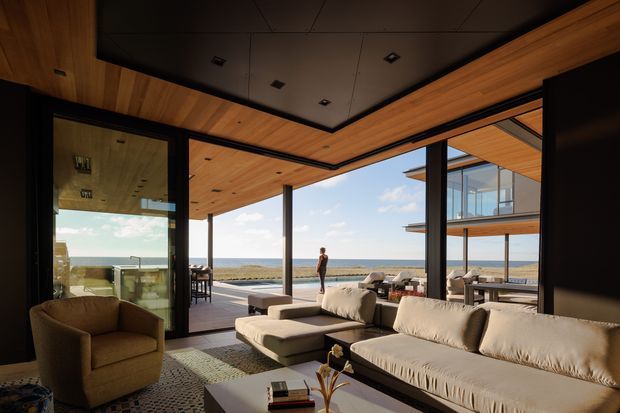 A sophisticated open-plan living room opens onto a broad outdoor terrace through extensive floor-to-ceiling glazing. The space features a large cream sectional sofa, armchair, low coffee table, dark structural posts, and warm timber-lined soffits surrounding a dark recessed ceiling. Beyond the terrace is a clear ocean or coastal horizon, with an adjacent upper-level glazed structure visible outside. The composition emphasises indoor-outdoor connection, expansive views, and refined contemporary detailing.