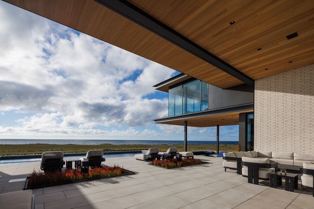 A sophisticated modern coastal terrace overlooking the ocean, framed by broad cantilevered roof planes with warm timber soffits. The outdoor living area features pale tiled paving, low lounge seating, sculptural chairs, built-in planted beds, slender dark columns, and expansive glazed walls. A light textured brick wall anchors the right side, while the open horizon and landscaped coastal setting create a strong indoor-outdoor connection.