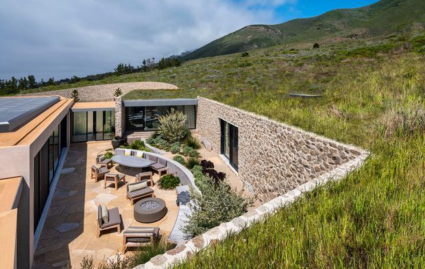 A contemporary hillside residence partially embedded into a sloping landscape, viewed from above. The image shows a sheltered central courtyard with outdoor lounge seating, a circular fire pit, paving, extensive floor-to-ceiling glazing, and warm-toned flat-roofed architectural forms. A substantial rough natural-stone retaining wall and planted green roof blend the building into the rugged mountain setting.