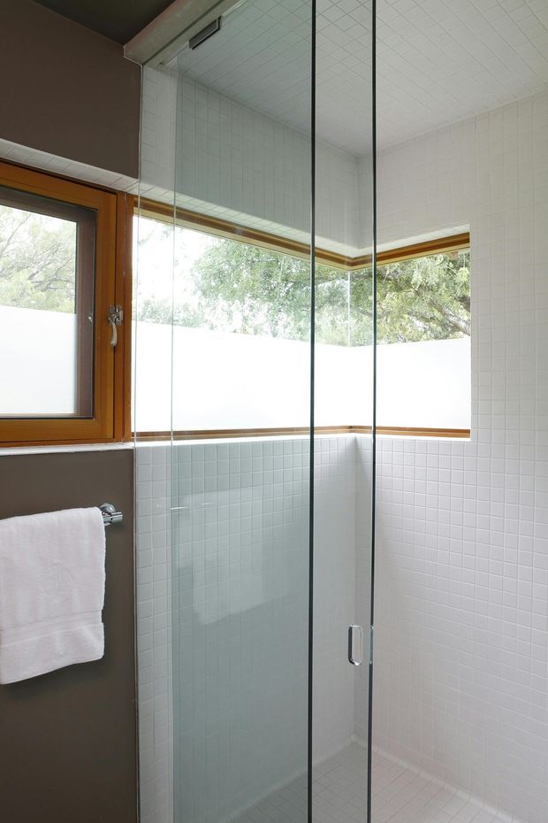 A bright, compact bathroom shower enclosure with clear glass panels and a frameless glass door. The shower walls and floor are covered in small white square tiles, while warm timber-framed windows wrap around the corner and bring in natural light. A white towel hangs on a chrome rail against a muted taupe wall.