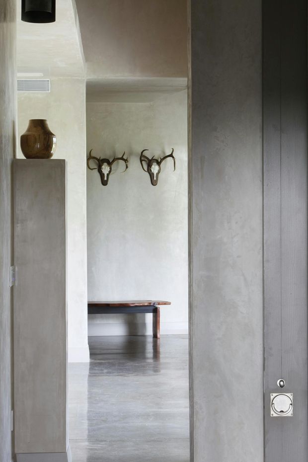 A narrow, moody view through a doorway into a minimalist interior. Pale textured plaster walls frame a polished grey concrete floor, with a simple dark timber bench in the background and two mounted antler sculptures on the wall. A tall pale fixed cabinet or pedestal with a ceramic vessel stands at left, while a dark vertically grooved door or panel with metal hardware occupies the right foreground. A black cylindrical pendant light is partially visible overhead.