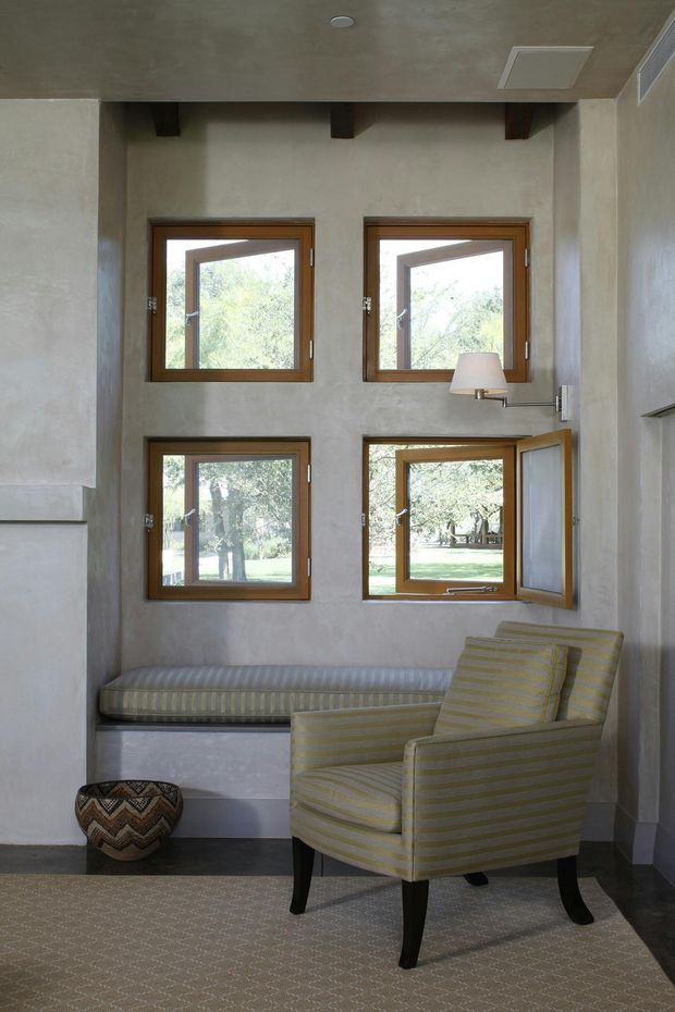 A compact, softly lit sitting nook with four timber-framed awning windows set into textured pale plaster walls. A built-in upholstered window seat runs beneath the lower windows, accompanied by a striped armchair, wall-mounted reading lamp, patterned footstool, and a low-pile neutral rug. Dark exposed ceiling beams and a muted, warm neutral palette add character to the otherwise simple space.