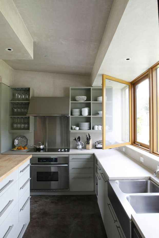 Compact contemporary galley kitchen with muted grey-green cabinetry, open shelving displaying glassware and crockery, stainless-steel appliances and splashback, pale stone worktops, and a deep stainless-steel double sink. Timber-framed awning windows bring in abundant natural light. The room has a simple plastered ceiling with a deep structural beam and dark flooring.