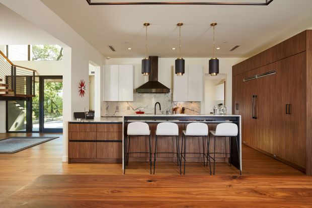 A warm, spacious contemporary kitchen viewed from the main living area, featuring a large island with four bar stools, white upper cabinetry, dark timber cabinetry and integrated appliances, a marble-look splashback, and three black pendant lights. Timber flooring continues through the space, with an open entry and staircase visible to the left.