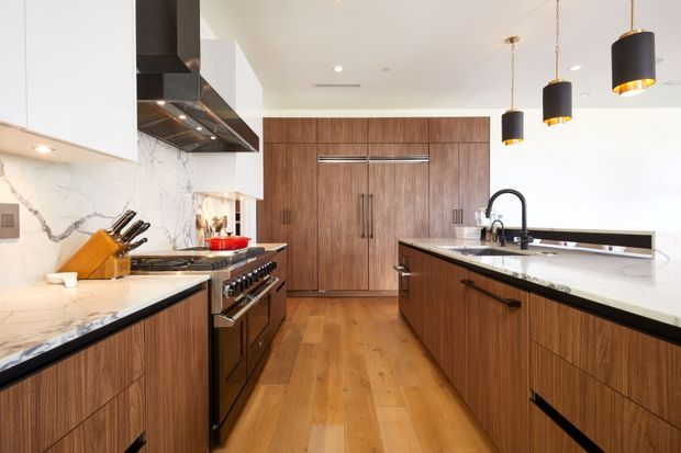 A bright, contemporary kitchen with warm vertical-grain timber cabinetry, white marble-look benchtops and splashback, a large island with an undermount sink and black gooseneck tap, and integrated stainless-steel cooking appliances. Three black pendant lights hang above the island, while full-height timber storage occupies the rear wall. Warm timber flooring and a clean, high-contrast palette create a refined, inviting space.