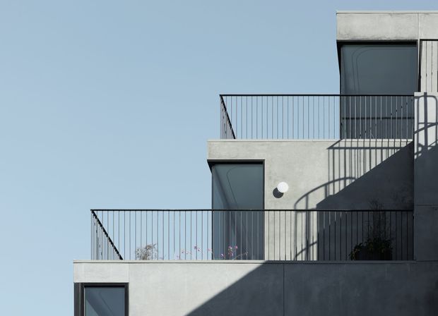 Minimalist multi-level concrete building facade with projecting terraces, dark metal balustrades, recessed dark-framed windows and strong angular sunlight and shadows. The image focuses on the exterior architecture against a clear blue sky.