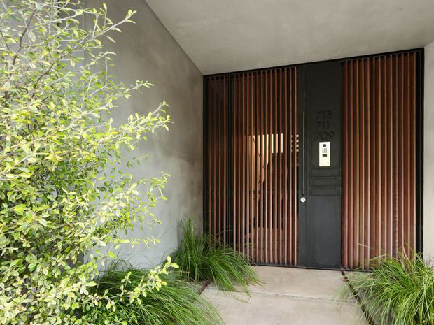 A contemporary residential entryway recessed beneath a concrete canopy. A dark door with an intercom is framed by vertical timber slats, with textured grey walls, a pale concrete path, and lush ornamental planting creating a layered, private entrance.