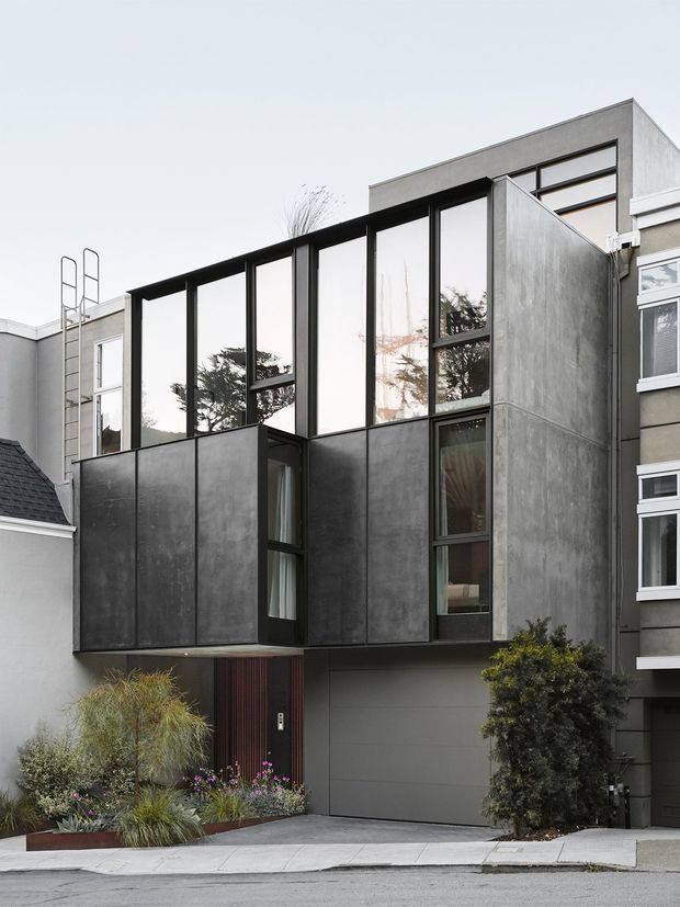 Street-facing exterior view of a contemporary multi-storey urban residence, featuring a projecting box-like addition clad in dark grey panels with tall black-framed glazing. The facade combines solid geometric volumes, narrow vertical windows, a garage door, and a landscaped entry garden, set among closely spaced neighbouring homes.
