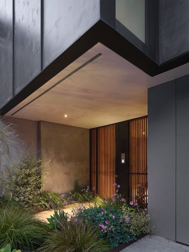 A moody contemporary residential entry courtyard viewed from outside. The sheltered entry features dark charcoal wall cladding, a warm smooth ceiling soffit with recessed lighting, a timber slatted security gate and door, and a textured brown-toned wall. Layered ornamental grasses and purple flowering plants soften the paved approach.