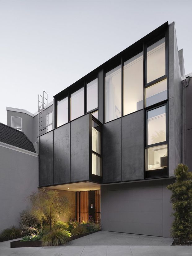 Contemporary multi-storey urban residence viewed from the street, with a dramatic cantilevered upper level, dark metal-clad facade, expansive black-framed glazing, and a recessed timber-lined entry above landscaped planting and a garage door. The home is integrated beside older neighbouring residential buildings.