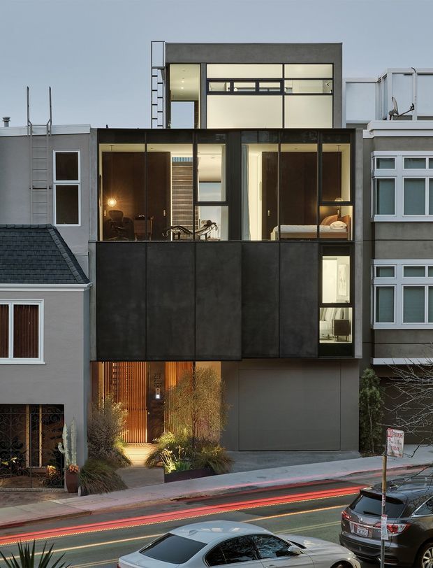 Exterior view of a narrow multi-storey urban residence inserted between neighbouring homes. The contemporary facade combines dark metal cladding, extensive floor-to-ceiling glazing, projecting box-like volumes, and a warm timber-lined entry landscaped with planting. Adjacent traditional residential buildings and a street frontage are visible.