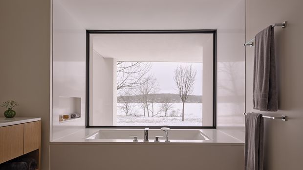 A refined contemporary bathroom with a built-in rectangular bathtub positioned beneath a large black-framed picture window overlooking a snowy landscape. The room has pale stone or tiled wall finishes, a warm wood floating vanity at left, minimalist chrome tapware, and twin wall-mounted towel rails with taupe towels. The composition is calm and softly lit, with a strong indoor-outdoor connection.
