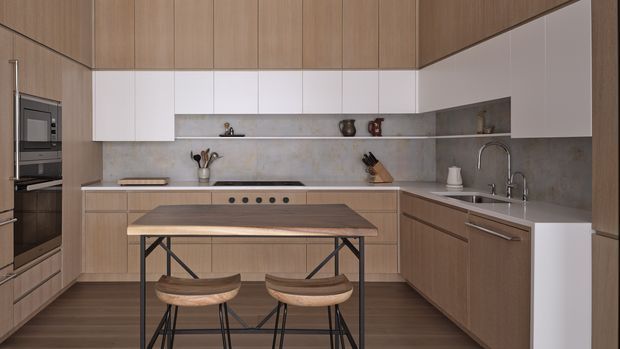 A contemporary galley-style kitchen with extensive warm wood cabinetry, white upper cabinets, and a large grey stone-look splashback and benchtop. A central timber island with dark metal legs is paired with two timber stools. Integrated appliances, a cooktop, sink, curved faucet, open shelving, and understated kitchenware complete the refined, modern fit-out.
