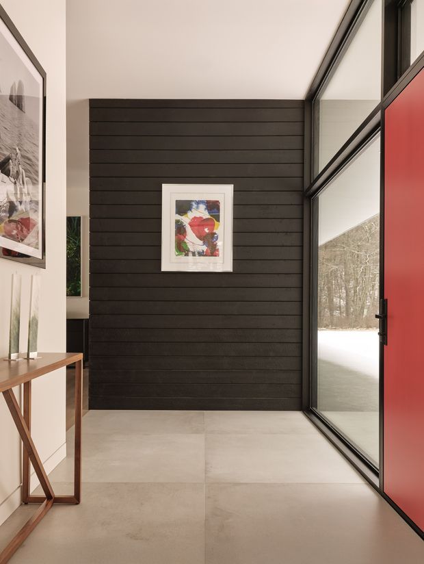 A refined residential entryway with a dramatic horizontal dark timber feature wall, large pale-format floor tiles, and full-height black-framed glazing beside a bold red pivot door. A slim timber console with decorative objects and framed artwork adds warmth, while the glazing looks onto a snowy wooded landscape.