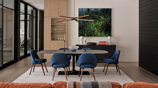 Contemporary dining area with a rectangular table surrounded by six blue upholstered chairs, positioned in front of a dark sideboard and built-in timber display cabinetry. Black-framed glazed doors line the left side, while a large wall-mounted artwork and sculptural linear pendant light provide focal points. Warm timber flooring, neutral walls, and a textured rug create a refined, inviting interior.