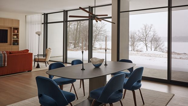 A modern dining area with a dark oval dining table surrounded by blue upholstered chairs, set beside expansive black-framed floor-to-ceiling glazing overlooking a snowy landscape and waterfront. The room also includes warm timber flooring, built-in timber cabinetry, a rust-coloured sofa, and a sculptural suspended timber-and-metal light fixture.