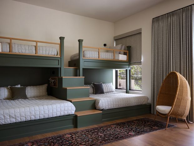 A well-designed bedroom featuring two built-in bunk-bed units with dark green painted cabinetry, integrated stairs, and quilted bedding. A large woven egg-shaped lounge chair sits beside full-height textured curtains, while a patterned rug and timber flooring add warmth. The room has soft neutral walls, compact windows, and a cozy, tailored built-in layout.
