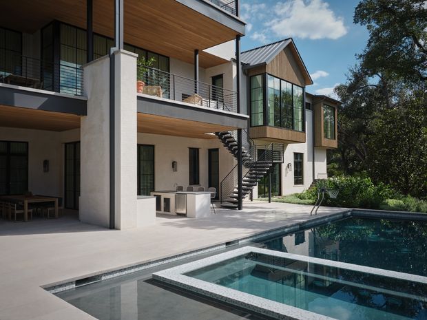 A contemporary multi-storey residence viewed from the pool terrace, featuring white rendered walls, extensive black-framed glazing, projecting balconies with horizontal metal balustrades, warm timber soffits, and a prominent pitched metal-roofed glazed volume. An exterior stair connects the levels, while the foreground contains a large swimming pool with integrated raised rectangular water features. Mature trees frame the property.