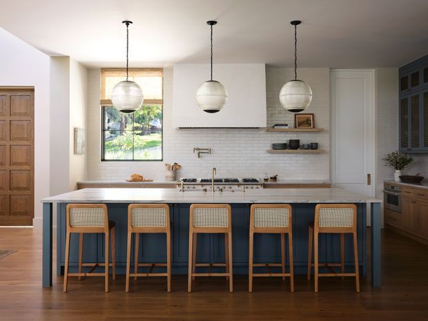 Bright, spacious contemporary kitchen with a large navy-blue island and five woven-seat timber bar stools. The room features warm timber flooring and cabinetry, white shaker-style and flat-panel joinery, a white subway-tile splashback, brass-toned hardware, globe pendant lights, and a rear door and window opening toward a garden. A timber entry door is visible on the left.