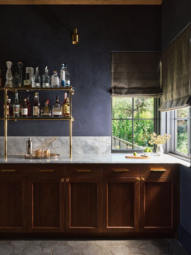 A richly detailed home bar with dark navy plaster walls, dark timber cabinetry, a pale veined stone benchtop and backsplash, brass shelving and hardware, and a marble-topped drinks counter. A corner window with Roman blinds brings in natural light and overlooks greenery. The floor features rounded patterned stone or ceramic tiles, creating a moody, refined atmosphere.