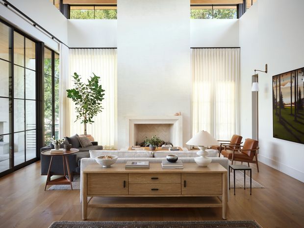A bright, sophisticated double-height living room with floor-to-ceiling black-framed glazing, clerestory windows, warm timber flooring, a central fireplace, and a large light-wood console table. The room is furnished with neutral sofas, sculptural lamps, a leather lounge chair, a small side table, and restrained artwork, creating a refined contemporary interior.