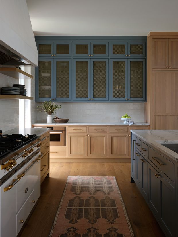 A refined kitchen with a warm, traditional-modern feel. The room features extensive light timber cabinetry, a contrasting dark blue-grey wall of upper cabinets with textured mesh or patterned glass fronts, white subway-tile backsplash, timber flooring, and a large dark island with a pale stone benchtop. A freestanding stainless-steel range with brass hardware sits to the left, while open timber shelving and decorative bowls add detail. The space is well lit with a balanced, warm-neutral palette.