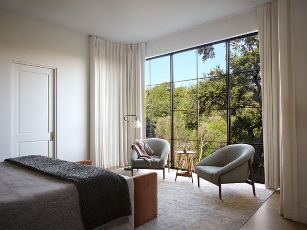 A calm, refined bedroom with a large floor-to-ceiling black-framed window overlooking trees. The room features a neatly made bed with a dark textured throw, two upholstered mid-century-style lounge chairs, a small timber side table, a slim floor lamp, full-height cream curtains, and a white paneled door. Soft neutral finishes and abundant natural light create an inviting atmosphere.