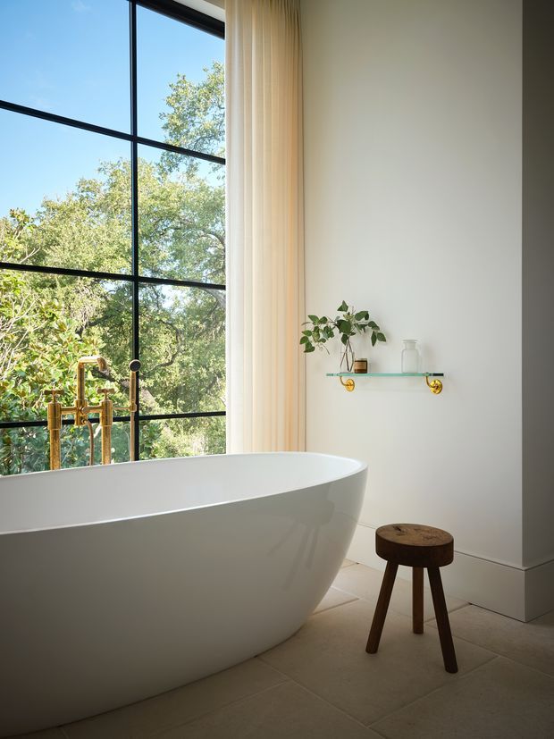 Bright, refined bathroom featuring a large white freestanding oval bathtub beside expansive black-framed glazing overlooking trees. A brushed-gold floor-mounted bath tap, pale curtains, light tiled flooring, and a simple glass shelf with minimal greenery complete the serene setting. A small dark timber stool adds contrast.