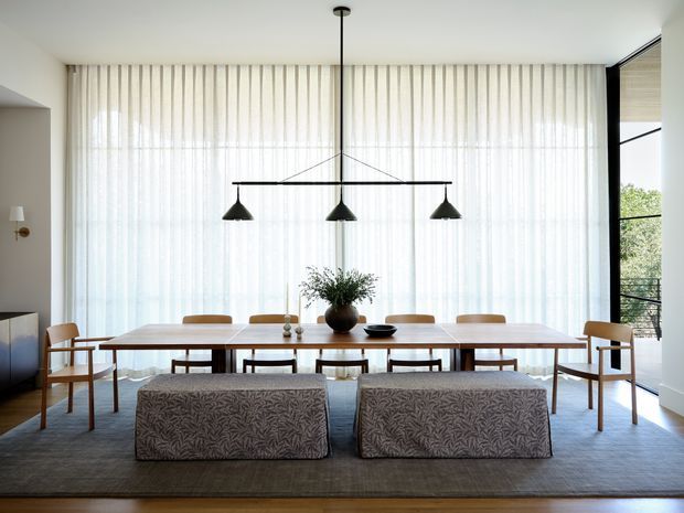Bright, spacious dining room with a long timber dining table and numerous wooden chairs, two upholstered bench seats in the foreground, and a linear three-light pendant suspended above the table. Full-height sheer curtains create a soft backdrop, with expansive glazing at the right and a restrained neutral palette of white, timber, grey, and black. A low sideboard and simple table styling complete the refined contemporary interior.