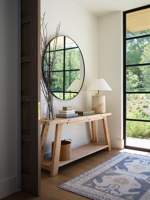 Bright, refined entryway featuring a light timber console table with open lower shelf, books, a ceramic table lamp, and decorative branches in a vase. A large round black-framed mirror reflects the garden and glazing, while a dark timber paneled door and full-height black-framed window/door provide contrast. The room has warm timber flooring, cream walls, and a muted patterned rug.