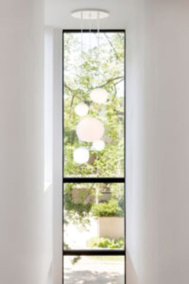 A narrow, tall interior window or glazed stairwell opening with black-framed divisions, extending across multiple levels. Bright daylight and leafy garden views are visible through the glazing, with round pendant lights hanging in front of the window. The surrounding interior is minimal, white, and softly lit.