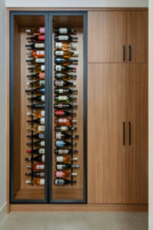 A tall built-in wine storage unit with two glass-front display sections holding numerous horizontally stored wine bottles, flanked by warm timber cabinetry with dark vertical handles. The cabinetry is set against a light neutral wall and floor, creating a clean, practical wine-storage feature.