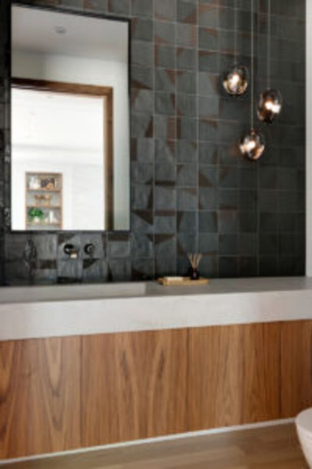 Contemporary bathroom vanity with a long white benchtop, warm timber floating cabinetry, and a dark textured tiled feature wall. A large frameless mirror reflects a bright adjacent space, while three spherical pendant lights provide a decorative accent above the vanity.