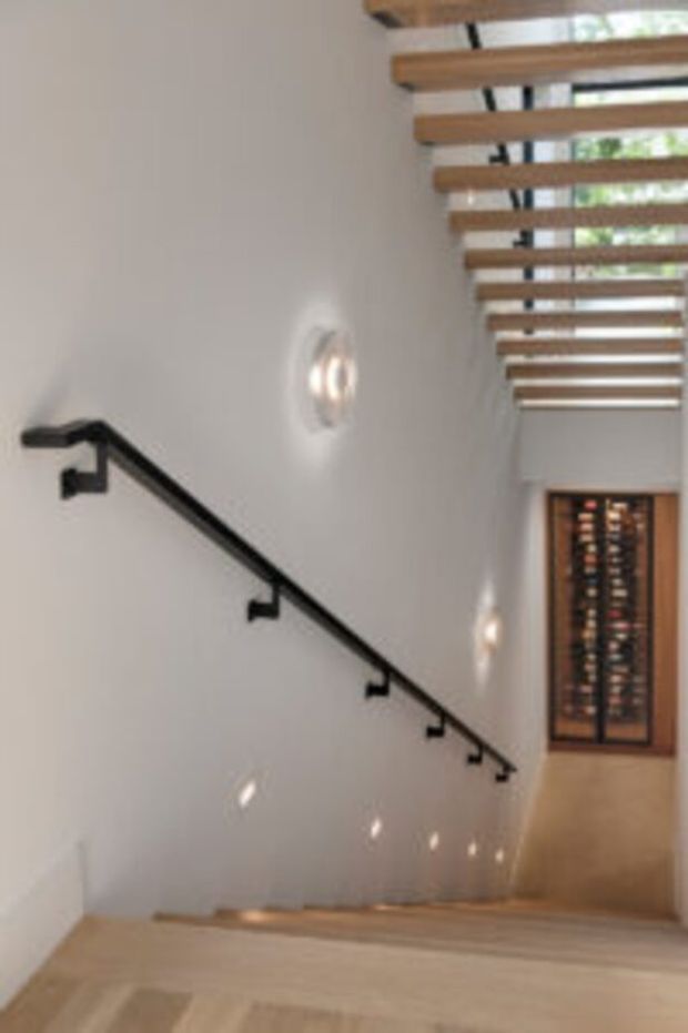 Interior view of a modern stairway descending toward a wood-framed glass door or feature at the far end. The stair has pale timber treads, smooth white walls, a slim black metal handrail, and small wall-mounted lights. Exposed timber ceiling battens create a strong linear rhythm overhead, with warm natural light entering through the upper glazing.