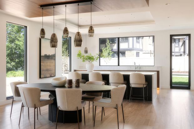Bright contemporary open-plan dining and kitchen interior with a large dining table and upholstered chairs in the foreground, a compact island or breakfast bar with stools, and extensive black-framed glazing. The room has pale timber flooring, light walls and ceiling, dark built-in cabinetry, and a row of woven pendant lights over the dining and kitchen areas. Garden and neighbouring buildings are visible through the windows and door.