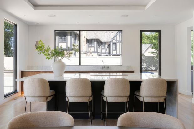 Bright contemporary kitchen with a large central island and four upholstered bar stools. The island has dark fixed cabinetry, a pale stone benchtop, and an integrated sink and tap. White walls and ceiling, light timber flooring, large black-framed windows and doors, and a sculptural white vase with greenery create an airy, refined interior. A neighbouring half-timbered building is visible through the glazing.