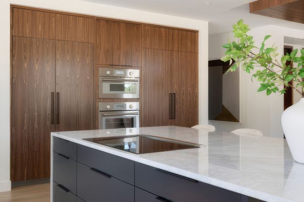 Contemporary kitchen featuring a large central island with a pale polished stone benchtop and dark integrated cooktop, dark charcoal drawer cabinetry, and a full wall of richly grained timber cabinetry housing stacked stainless-steel ovens. The room has white walls, recessed lighting, a partial timber ceiling detail, and a large white vase with greenery. A reflective surface or opening at the right provides a glimpse of adjacent space.