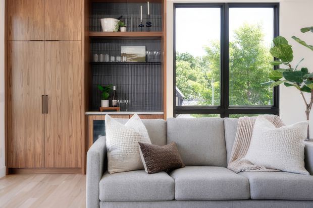 A bright, refined living room with a large light-grey sectional sofa in the foreground, integrated warm timber cabinetry and open shelving to the left, and a black vertically ribbed feature backing behind the shelves. A large black-framed window provides natural light and views of greenery, while textured neutral cushions, a woven throw, and indoor plants add softness and warmth.
